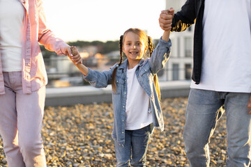 Smiling girl with parents holding hands at sunset, enjoying family time outdoors. Concept of happiness, unity, and childhood.