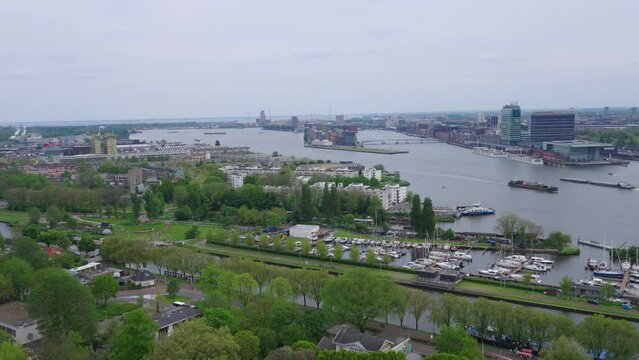 Western aerial view of north Amsterdam, with lush greenery, a canal, yacht docks, buildings and tanker ships in the IJ river, on a cloudy spring day 