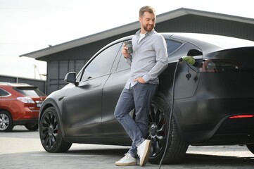 handsome bearded man in casual wear, standing at the charging station and charger for an electric car. Eco electric car concept