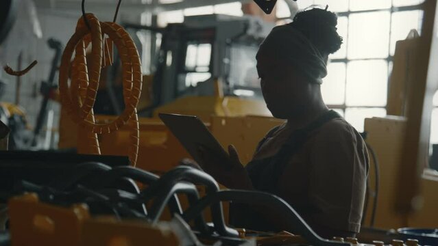 Young female industrial worker checking machine parts and typing on digital tablet while doing plant inspection