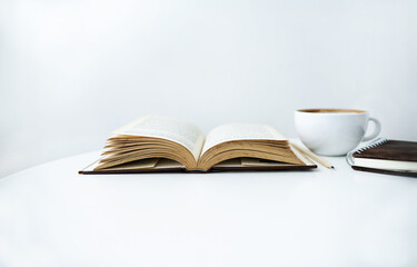 Close-up shot, white coffee cup and note book with open book on white table and white wall on background