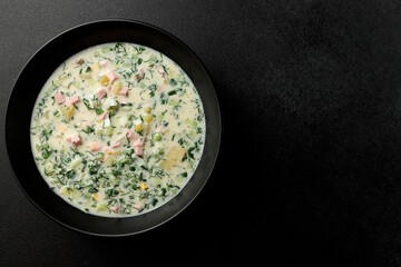 Cold soup with fresh cucumbers, sausage, broth and sour cream in a black plate on a black background. A traditional dish is okroshka