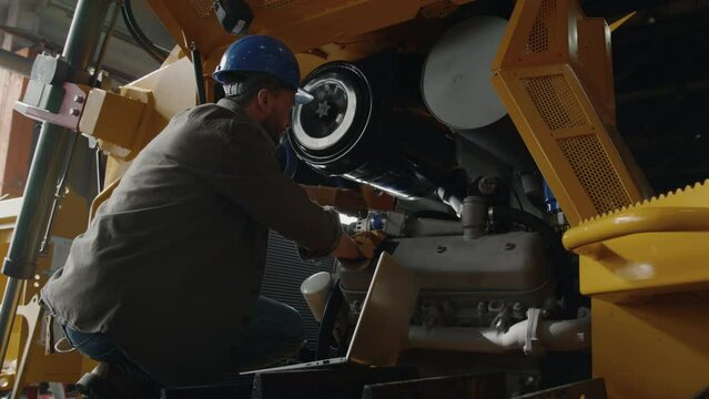 Technician in hardhat and workwear maintaining tractor with screwdriver and using laptop while working at industrial plant