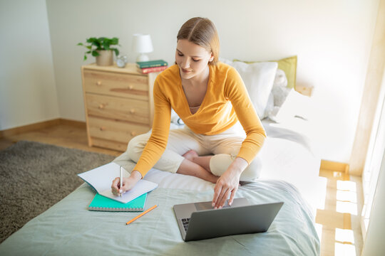 A teen girl sits on her bed in the morning, working on a laptop and writing in a notebook. She appears focused and productive, surrounded by a neat and cozy bedroom setting with natural sunlight