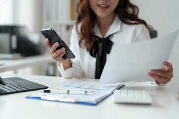 Happy Asian businesswoman in office while using smartphone to contact customer, online video call on documents, business finance, marketing, company taxes. Online business contact.
