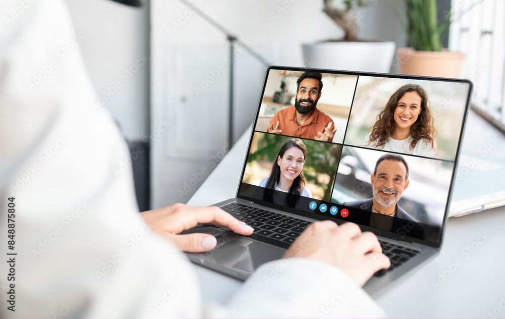 Poster Lady sitting at a laptop is engaged in a video conference with a group of people displayed on the screen. The individual appears focused as they interact digitally with their remote team.