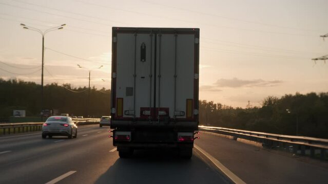White cargo truck van with freight semi-trailer driving along highway in evening time at sunset, rear view.