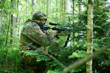 Hunter with rifle wearing camouflage suit sitting in ambush. Camouflaged sniper in the forest in...