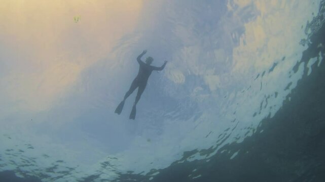 A woman snorkeler floats in the water, silhouetted against the sunset sky, while bubbles rise from below. Filmed from underwater straight up.