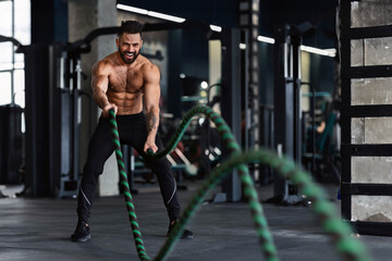 Fototapeta premium Crossfit training. Naked muscular man exercising with battle ropes at gym, free space