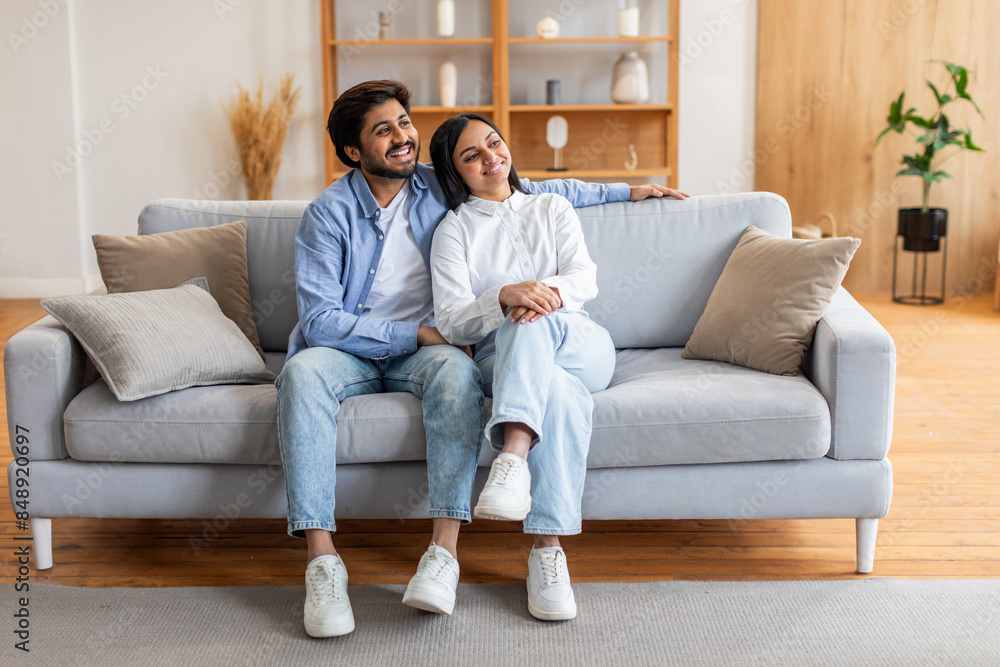 Wall mural Indian man and woman are seated on a beige couch in a living room. They appear to be engaged in conversation or watching TV, looking at copy space