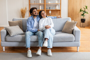 Indian man and woman are seated on a beige couch in a living room. They appear to be engaged in...
