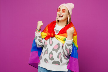 Young happy lesbian woman wrapped in rainbow flags wear pink animal clothes do winner gesture celebrate clenching fists isolated on plain purple background. Pride day June month love LGBTQ concept.
