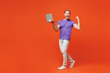 Full body young smiling happy IT man he wear purple t-shirt casual clothes hold use work on laptop pc computer do winner gesture isolated on red orange background studio portrait. Lifestyle concept