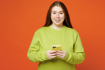 Young surprised fun fat ginger chubby plus size woman wear green sweatshirt casual clothes hold use mobile cell phone isolated on red orange background studio portrait Lifestyle body positive concept