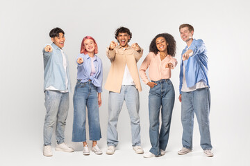 Obraz premium A group of five diverse friends, all smiling and pointing at the camera, are standing in front of a white background in a studio setting.