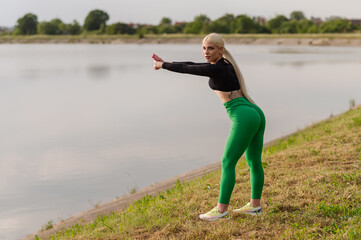 A girl in a sports outfit is warming up or doing exercises in a park on the shore of a lake