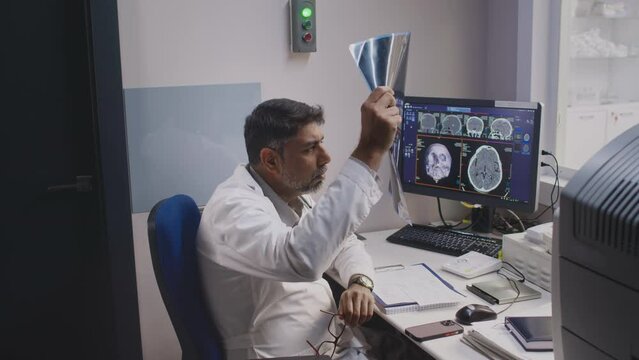 Medium shot of mature male radiologist wearing white lab coat and glasses sitting in front of computer examining Xray images in MRI room and detecting diagnosis
