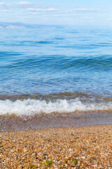 Natural background of clean gravel beach with transparent pure blue water with waves and a ridge of mountains on horizon on sunny clear day. Siberian Baikal Lake. Beautiful landscape of Olkhon Island 
