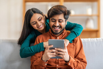 Indian man and woman are seated on a couch, both focused on a tablet screen. They appear engaged as they interact with the device, possibly watching a video or scrolling through information
