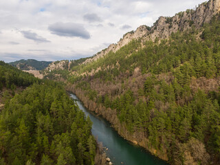 A river running through the centre of a valley, surrounded by pine trees. Cloudy sky.