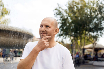 Bald man in white t-shirt touching chin while looking thoughtful near fountain