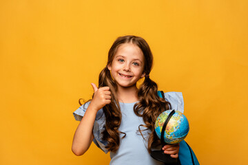 Showing thumbs up. Happy first year schoolgirl girl in a blue t-shirt and jeans on a yellow background with a globe in her hands and backpack. Copy space. Back to school concept.