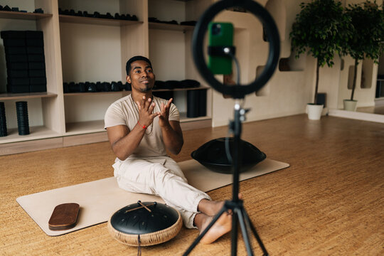 Focused African male blogger instructor sitting on yoga mat with Sadhu Board and glucophone, talking about practice in front of mobile phone with ring light while filming class for fitness channel. - Powered by Adobe