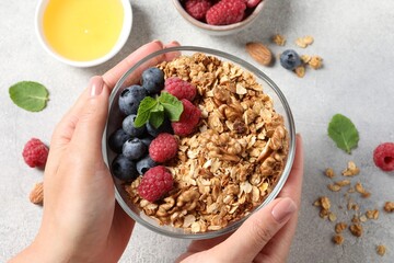 Woman holding bowl of tasty granola with berries, nuts and mint at grey table, top view