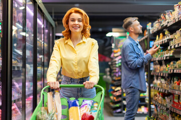 Fototapeta premium Cheerful lady doing shopping, buying products in supermarket, pushing trolley cart and smiling at camera