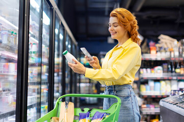Cheerful European woman scanning qr code of daily product using smartphone, standing near refrigerator. Concept of shopping and consumerism.