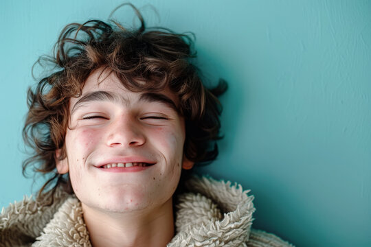 retrato de un chico joven adolescente sonriendo con ojos cerrados, pelo ondulado y cazadora de borreguito sobre fondo azul turquesa