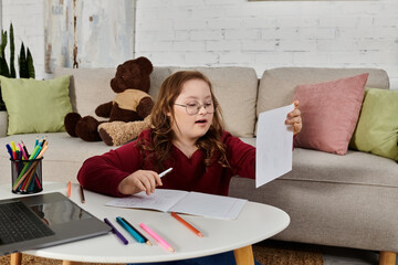 A little girl with Down syndrome sits at a table with art supplies, engrossed in her work.