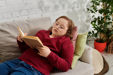 A little girl with Down syndrome relaxes on a couch at home, engrossed in a book.