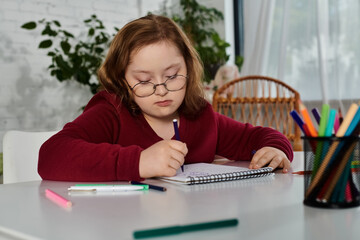 A little girl with Down syndrome sits at a table, intently drawing in her notebook.