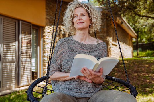 Smiling middle aged good-looking blond curly haired woman, reading a book in hammock in the garden during spring, summer.