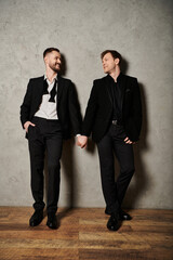 Two men in black suits stand close, smiling and holding hands.