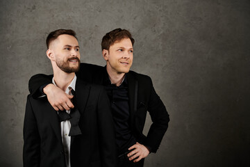 Two men in elegant suits pose for a photograph against a grey backdrop.