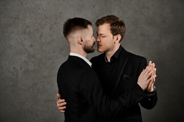 Two men in black suits stand close together, embracing and looking lovingly into each others eyes.
