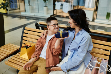 A mother and son with Down syndrome sit on a bench in a shopping mall, sharing a gift and smiling at each other.