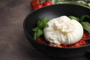 Delicious burrata cheese, tomatoes and basil in bowl on brown table, closeup