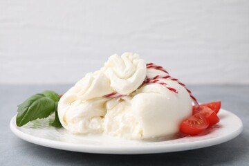 Delicious fresh burrata cheese with basil leaves and tomato on grey table, closeup