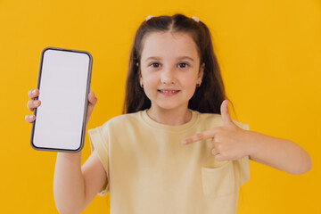 Smiling Caucasian kid girl against yellow wall Mock up copy space. Pointing index finger on mobile phone with blank empty screen