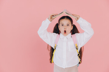 Cute little girl with open mouth in school uniform with backpack holding open book above head on...