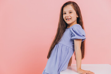 Little cute happy girl in blue dress posing in studio near white cube on pink background