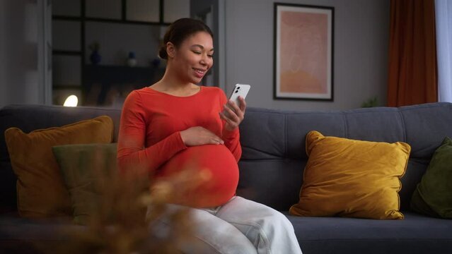 African American woman female pregnant girl at home pregnancy lady future mother at home relaxing stroking belly using mobile phone app chatting laughing smiling smartphone social media web device fun