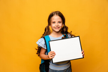 White banner. Little child schoolgirl in blue shirt on yellow background holding white blank paper. Young smiling girl show blank board. Copy space. Mock up.