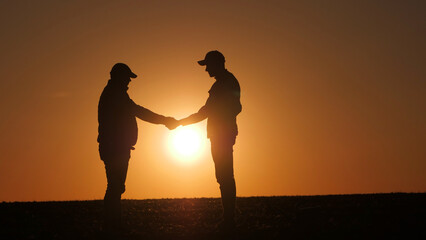 Against the setting sun over the field, the silhouettes of two farmers shake hands.