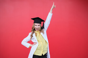 Young blond female in medical uniform and graduation cap with funny emotions and in a good mood. Standing on red background, happy to finish studying in healthcare or veterinary. High quality photo