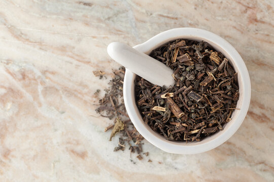 Top-view of Dry Organic Bhringraj (Eclipta prostrata) leaves, in white ceramic mortar and pestle, on a marble background.